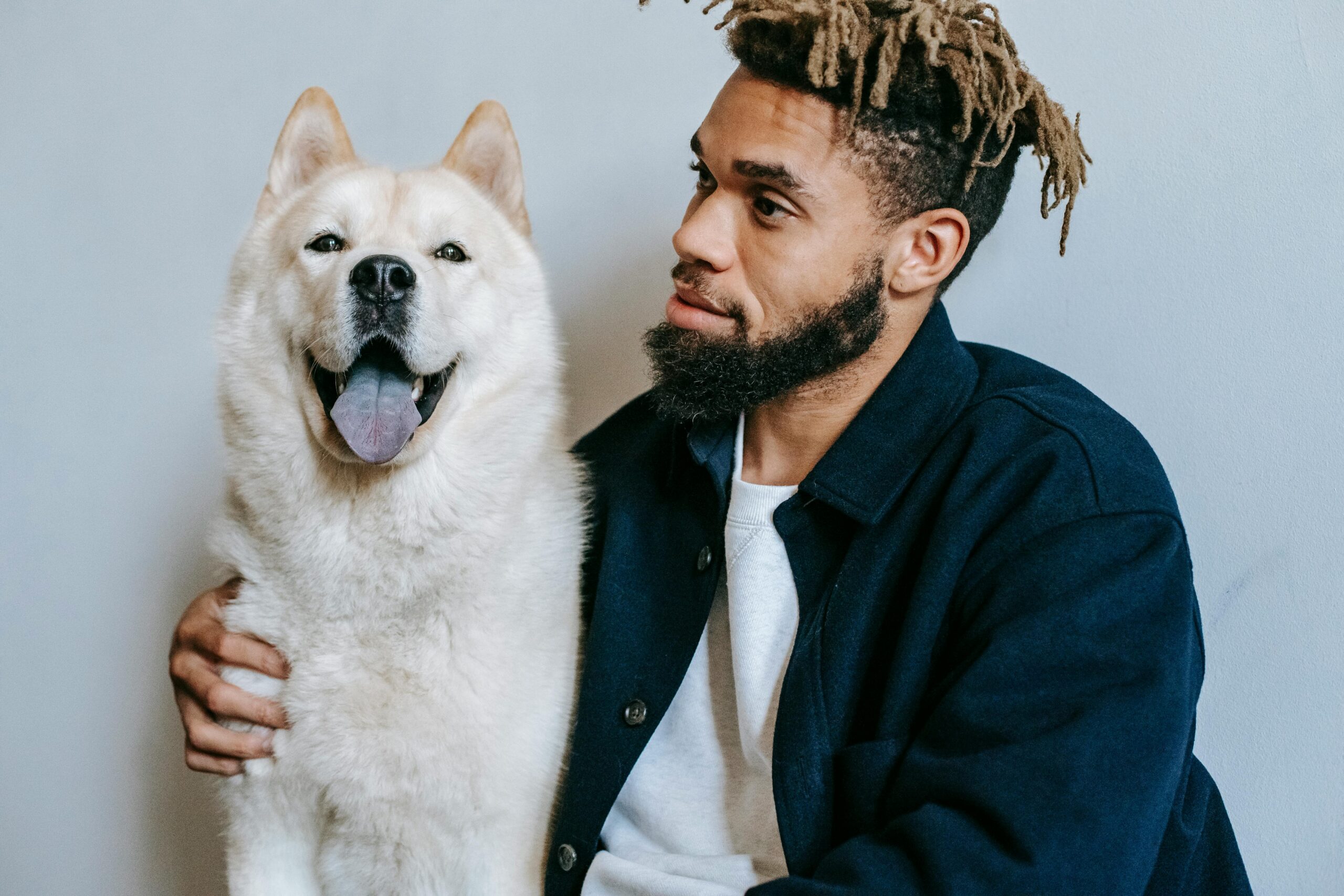 A young man with dreadlocks sitting indoors, accompanied by a cheerful Akita Inu dog.